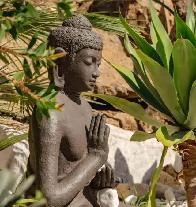 Figura decorativa de Buda en un jardín rodeado de plantas y piedras ornamentales.